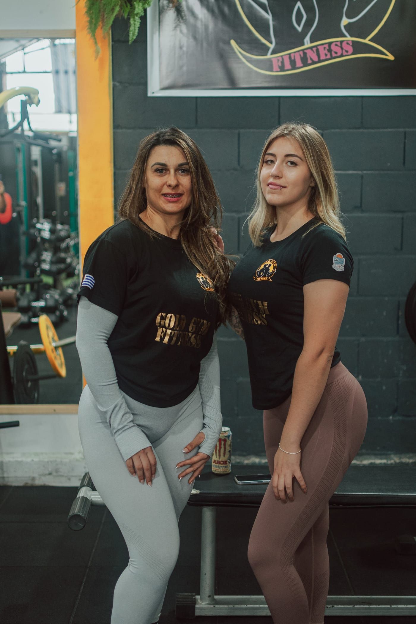 Dos mujeres con la remera de Gold Gym, frente a la pared del logo